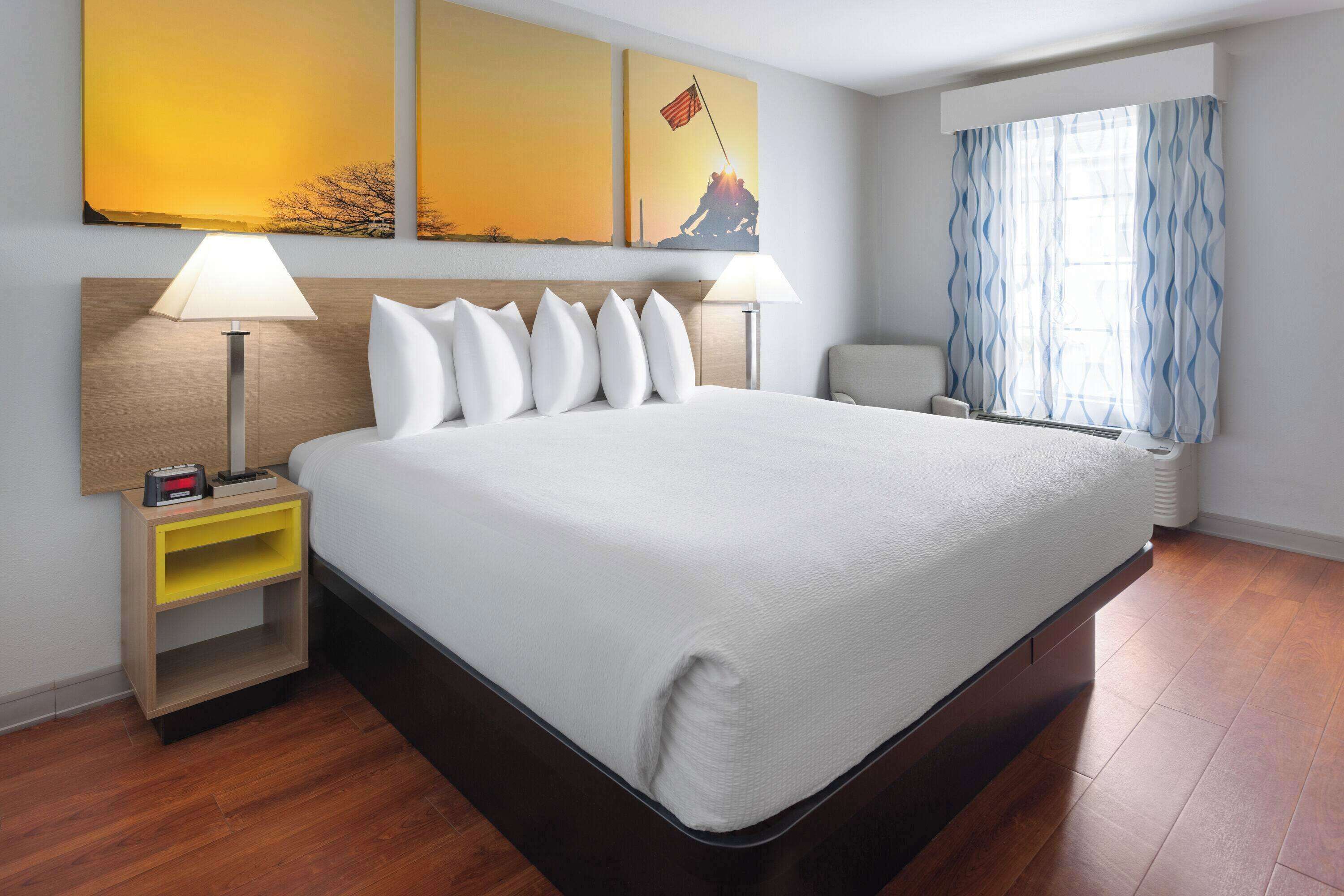 A hotel room with a large bed, two bedside tables, a nightstand, a window with curtains, and artwork on the wall.
