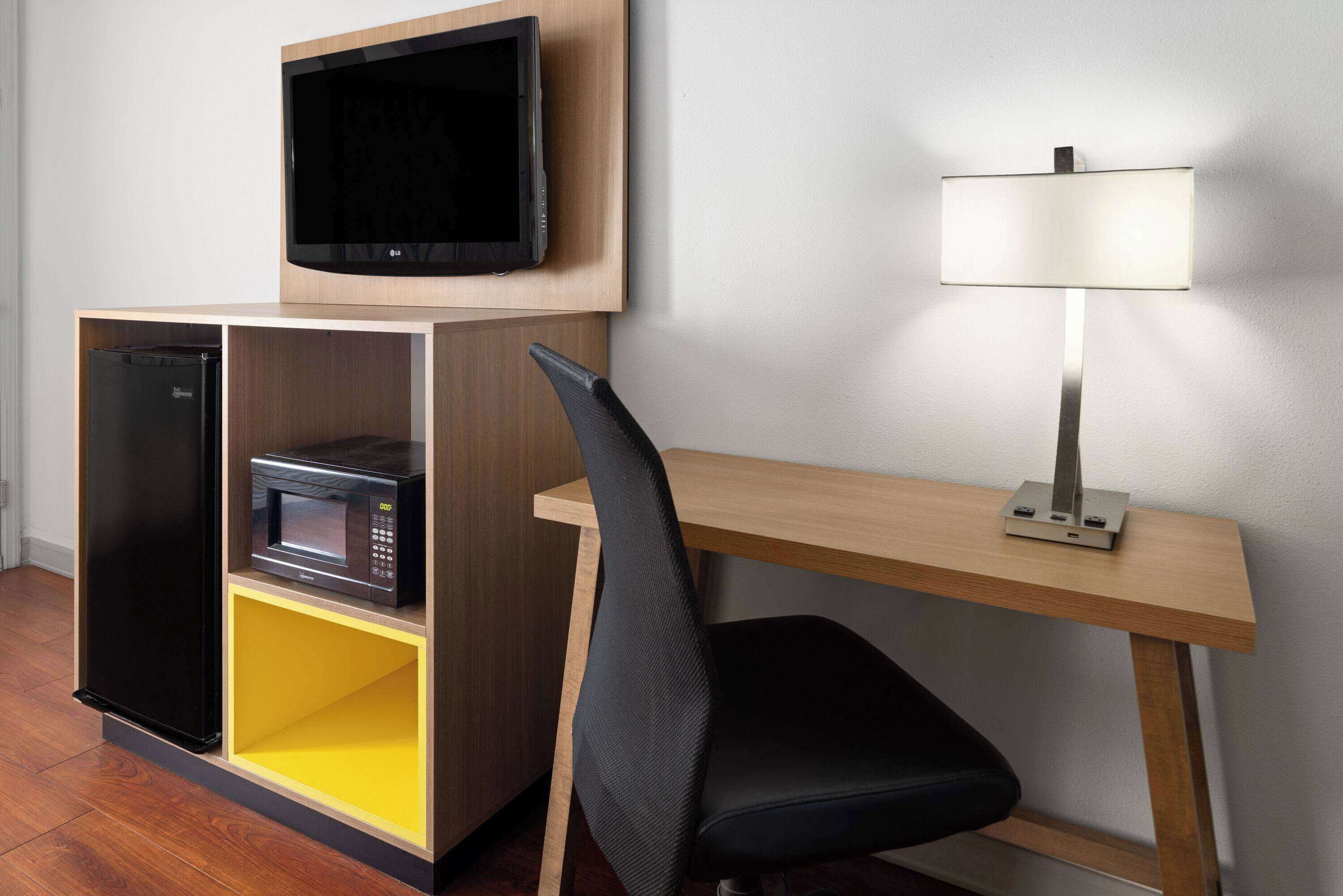 Una habitación de hotel pequeña con un escritorio de madera, una silla de oficina negra, un televisor de pantalla plana, un microondas y una lámpara.