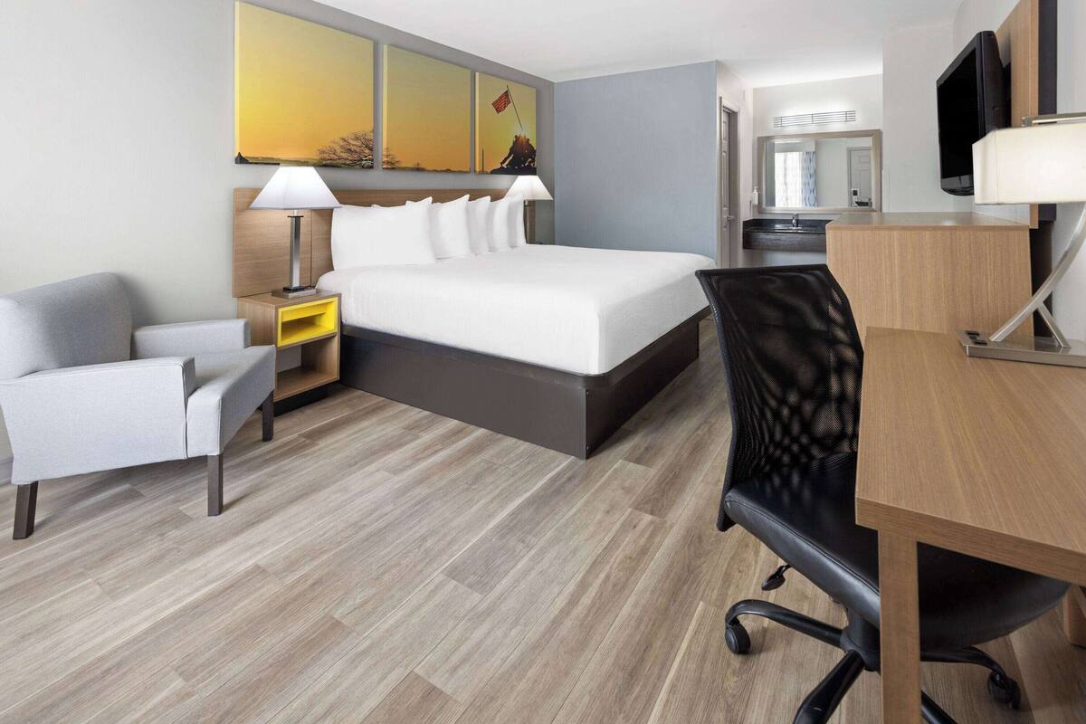 Quarto de hotel com cama, escrivaninha, cadeira, mesa de cabeceira e televisão.
