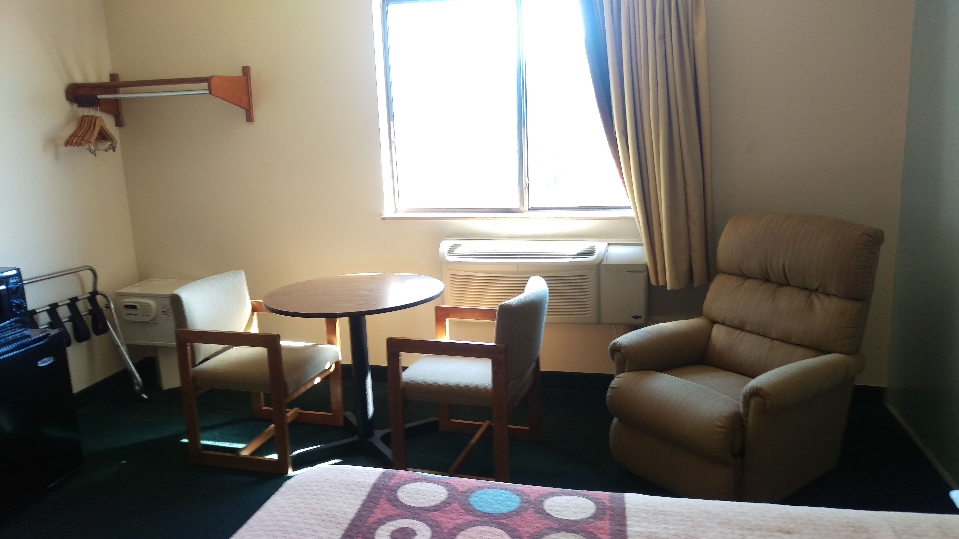 Een hotelkamer met een bed, een ronde tafel met twee stoelen, een leren fauteuil, een raam met gordijnen, een airconditioner en een kapstok met hangers.