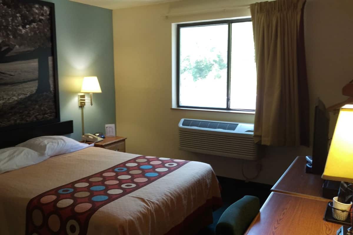 Quarto de hotel com cama, escrivaninha, luminária, janela com cortinas, ar-condicionado e um quadro na parede.