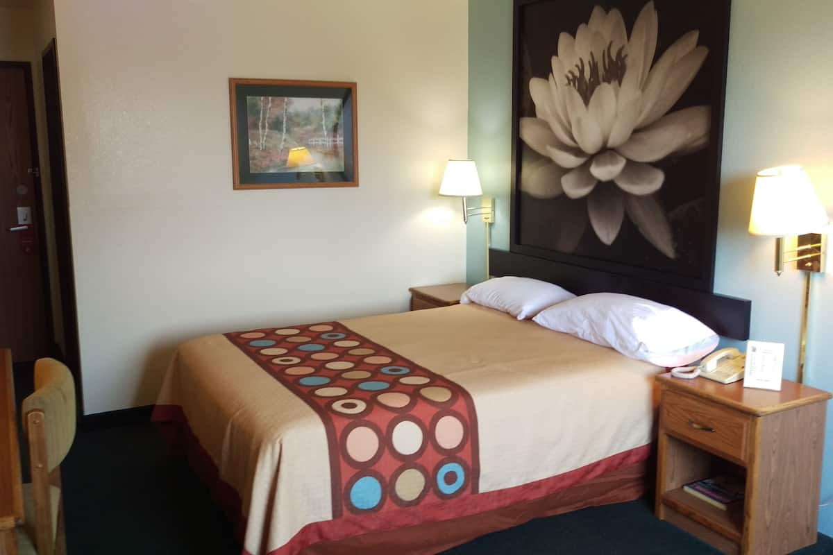 Quarto de hotel com cama, duas luminárias de cabeceira, mesa de cabeceira com telefone e luminária, uma cadeira de madeira, um quadro na parede e uma porta.