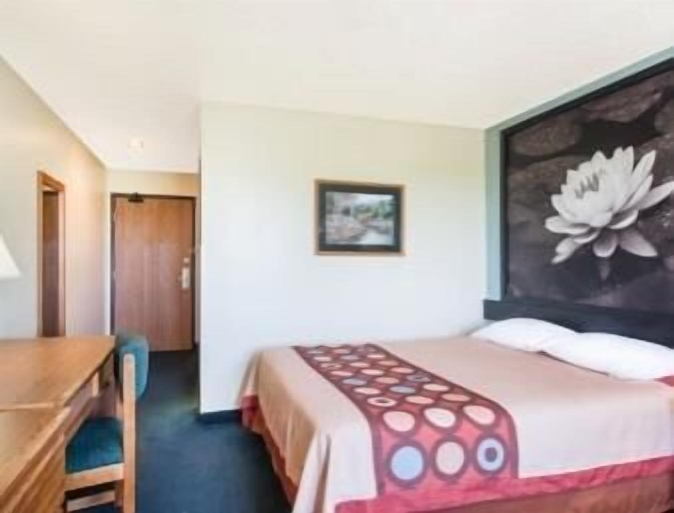 A hotel room with a bed, a desk, and a chair. There is a painting on the wall and a door in the background.