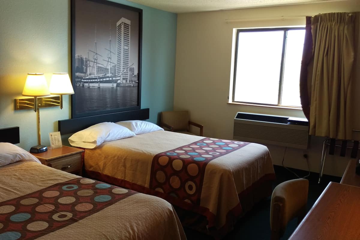 Quarto de hotel com duas camas, uma mesa de cabeceira com abajur, uma cadeira, uma janela com cortinas e uma pintura de paisagem urbana na parede.