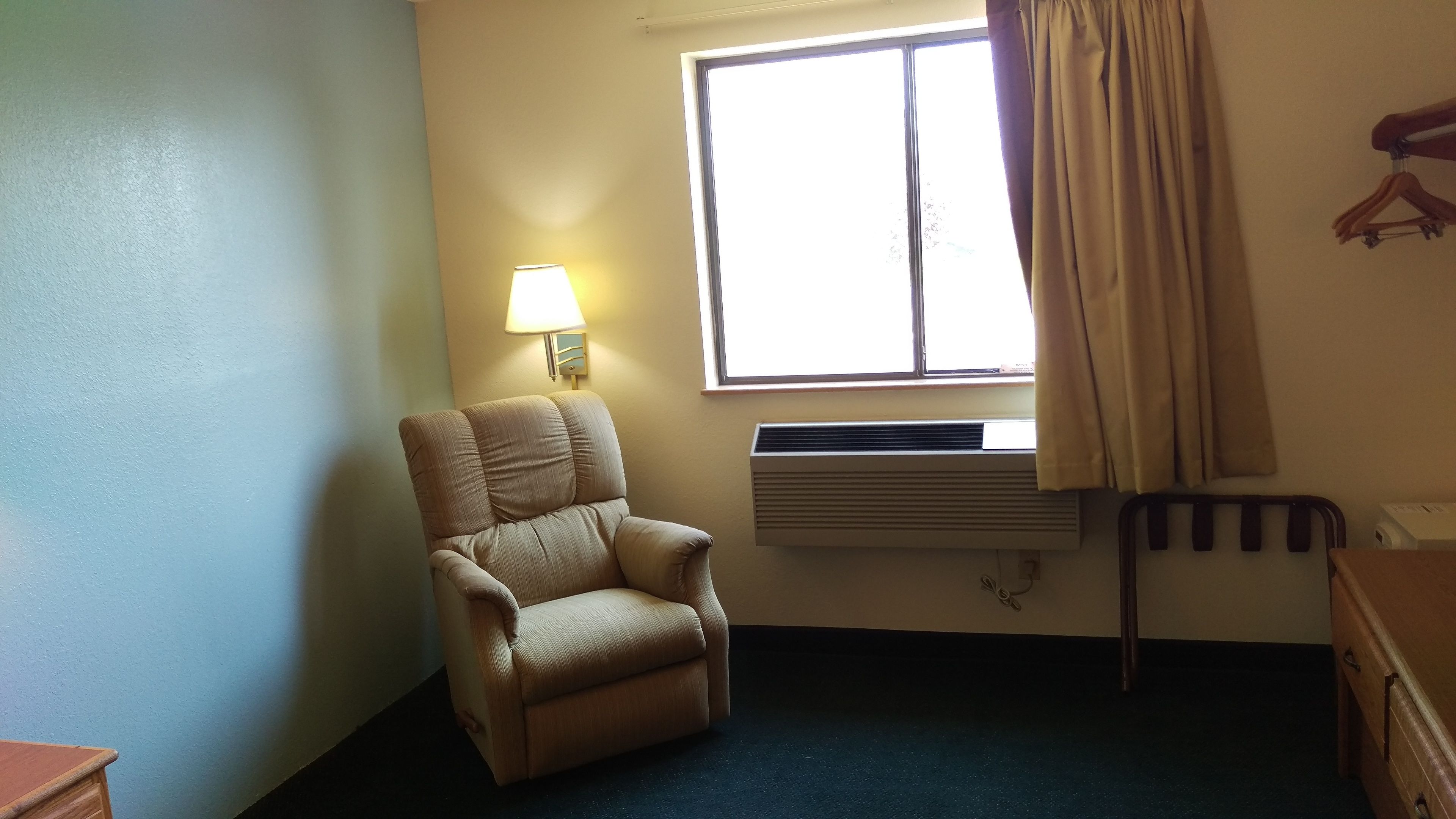 Una habitación con un sillón individual color beige, una ventana con cortinas, un aire acondicionado de pared y un aparador de madera.