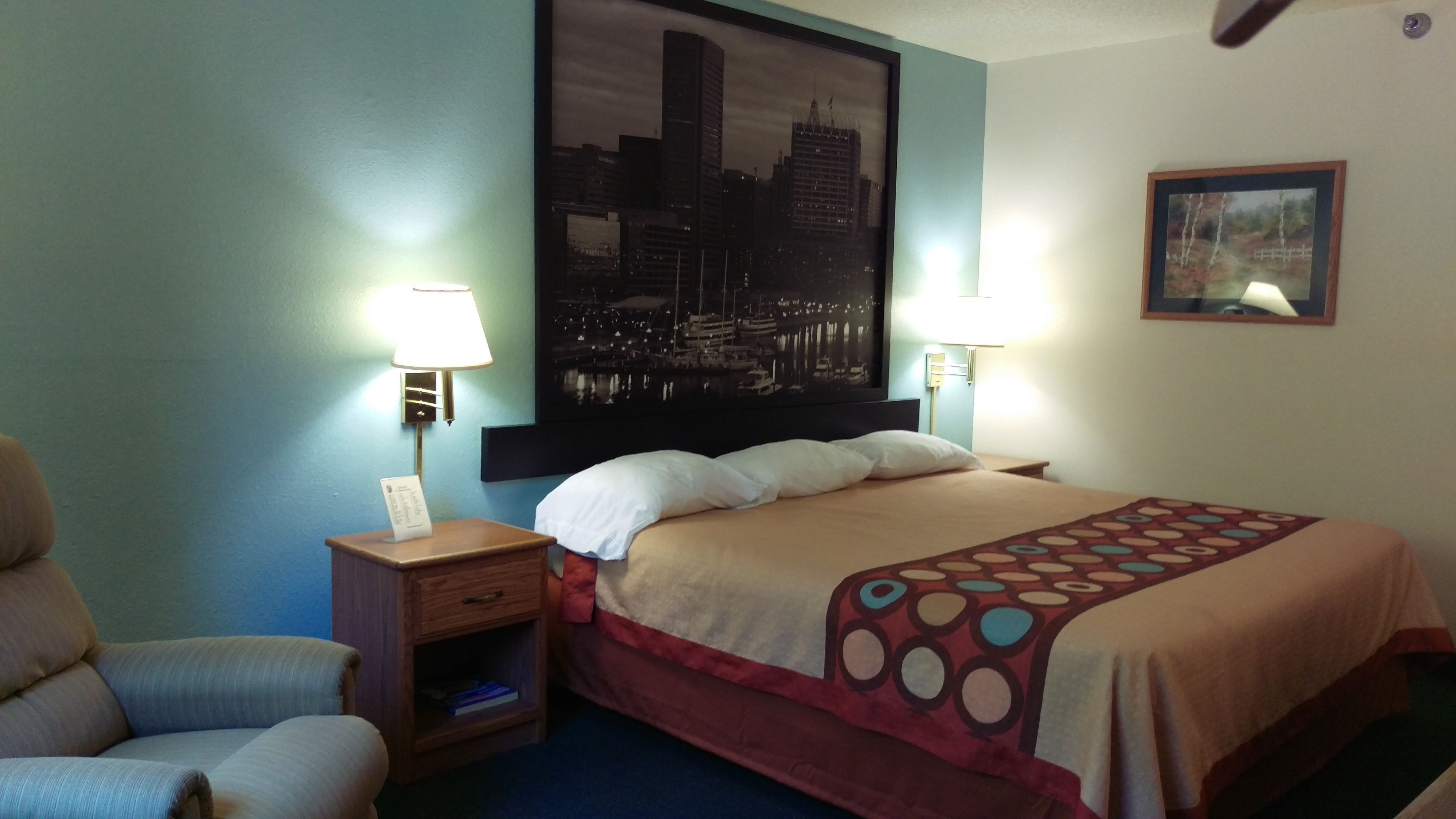 A hotel room with a bed, two bedside lamps, a nightstand, a chair, a cityscape painting on the wall, and a framed picture.