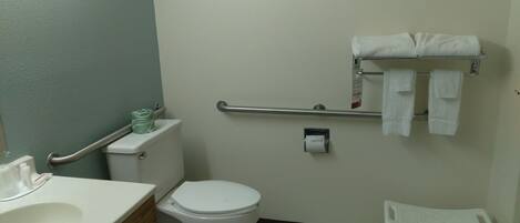 A bathroom with a toilet, a handrail, a sink, a small white chair, and a towel rack.