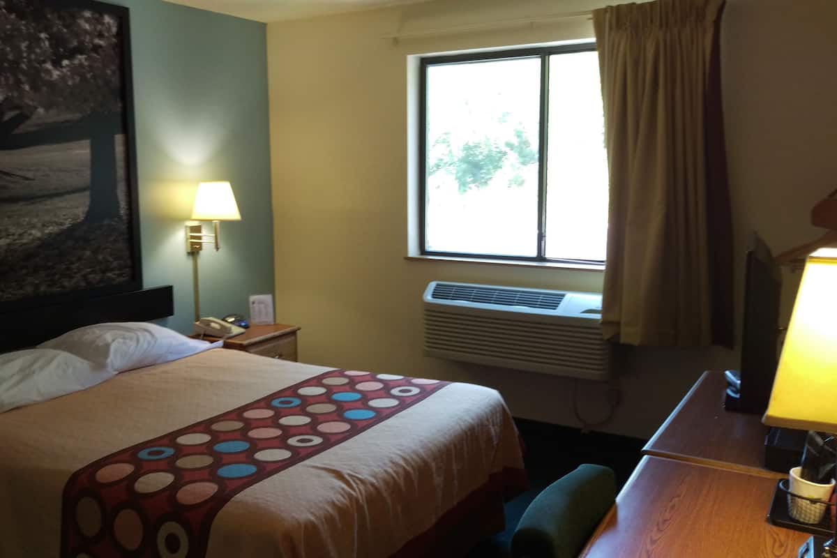 Quarto de hotel com cama, escrivaninha com luminária, janela com cortinas, ar condicionado e um quadro na parede.