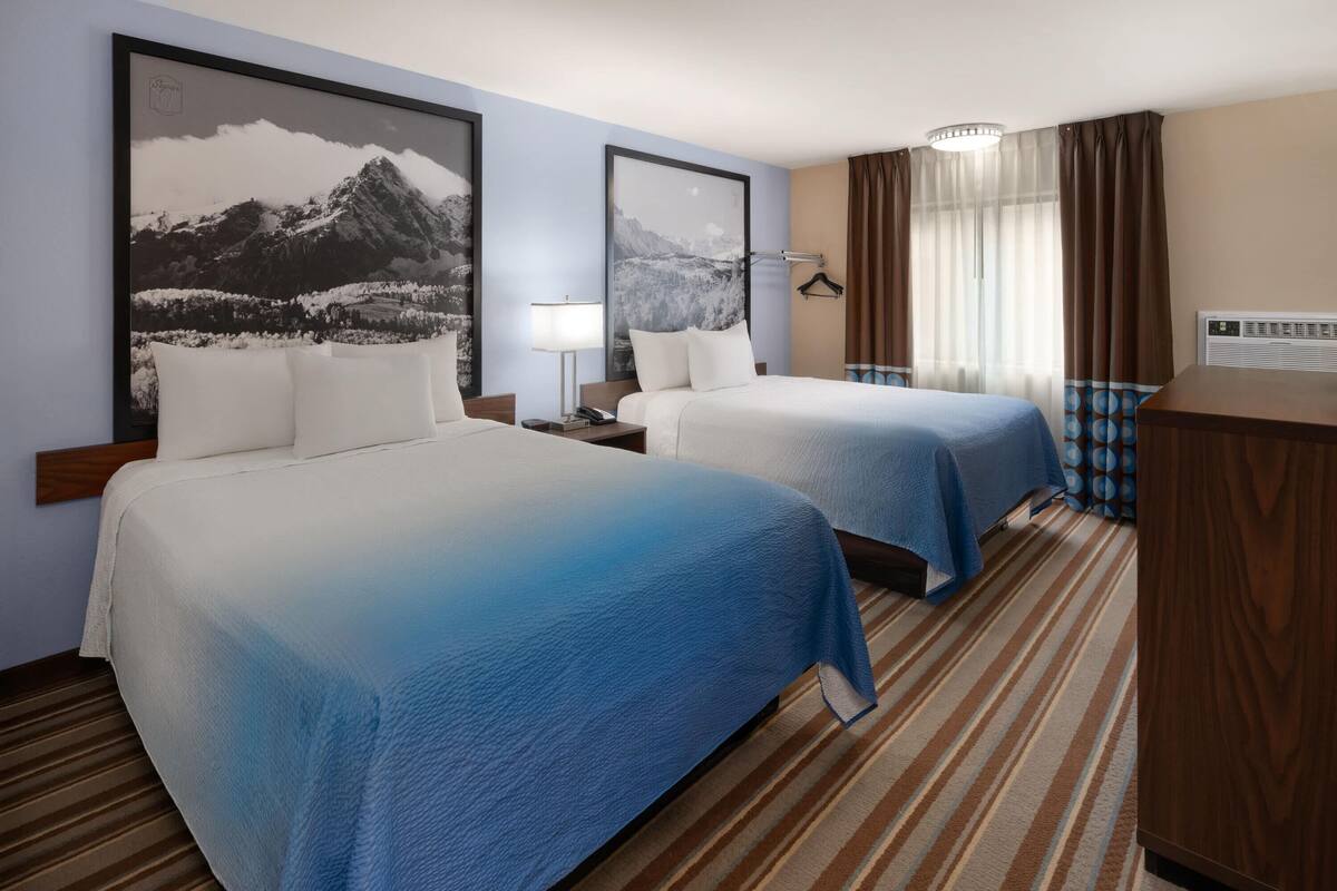 A hotel room with two beds, a desk, a lamp, a picture of mountains, a window with curtains, and a wall-mounted air conditioner.