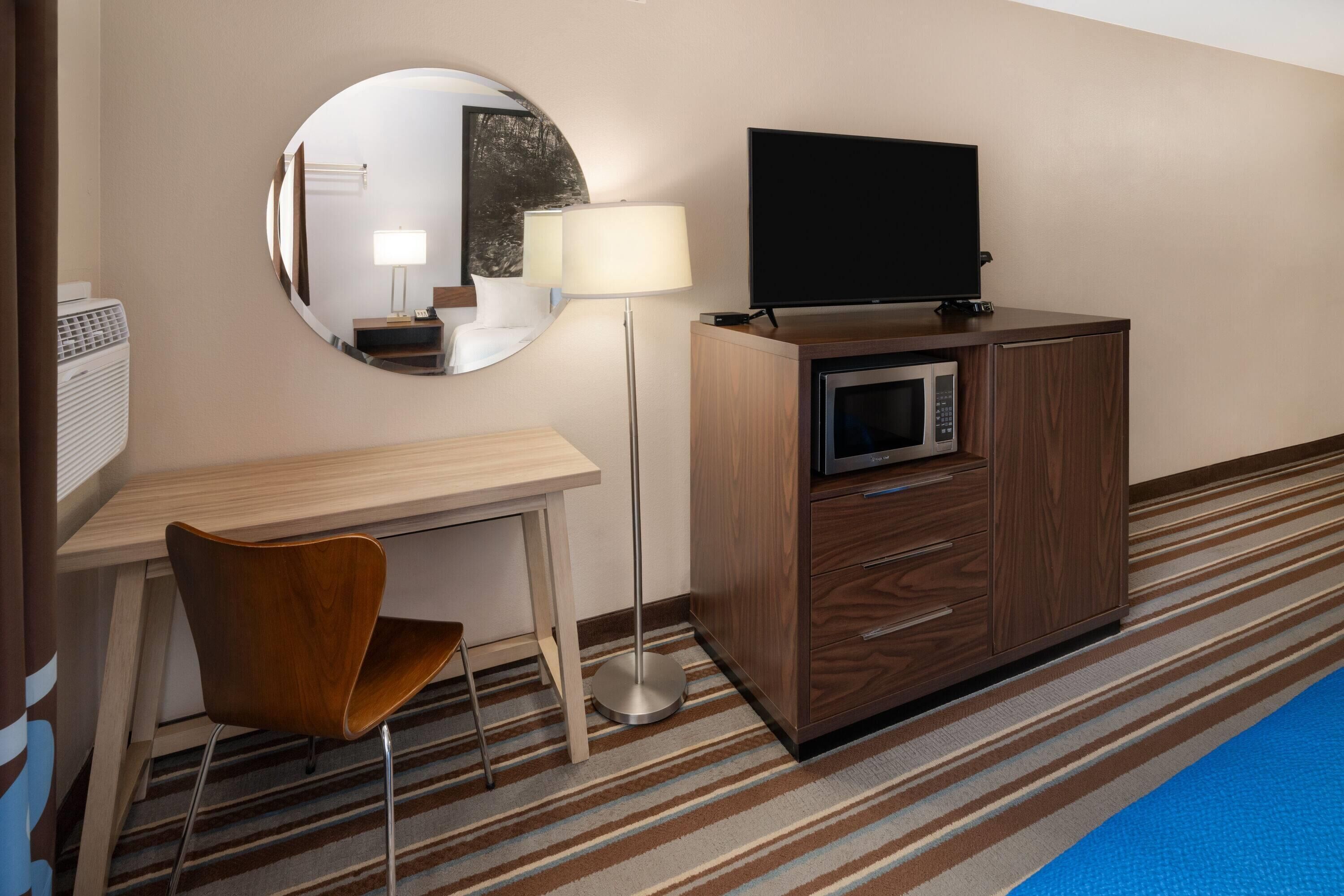 Habitación de hotel con escritorio de madera, una silla, televisor sobre un mueble, microondas, espejo y alfombra a rayas.