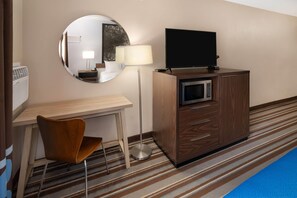 Una camera d'albergo con una scrivania in legno, una sedia, un televisore su un mobile, un forno a microonde, uno specchio e un tappeto a righe.