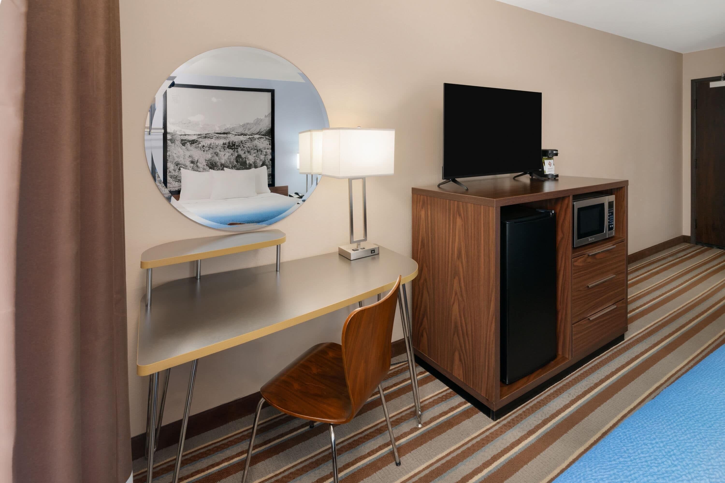 A hotel room with a bed, a desk with a chair, a television, a microwave, and a large mirror.