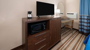 A hotel room with a wooden cabinet, a flat-screen TV, a microwave, a small table with a chair, a mirror, an air conditioning unit, and a striped carpet.