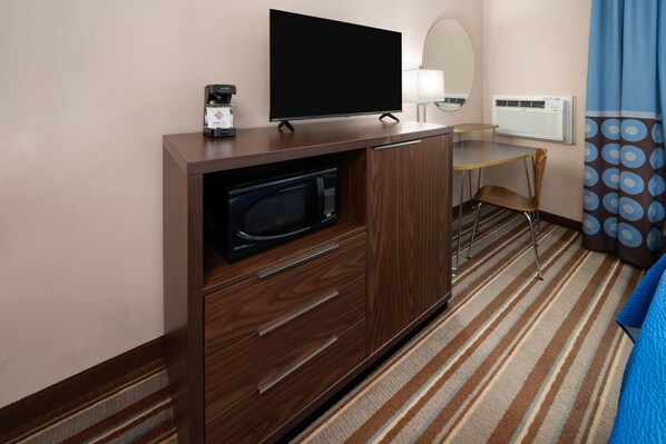 Camera d'hotel con un armadio in legno, una TV a schermo piatto, un forno a microonde, un piccolo tavolo con una sedia, uno specchio, un condizionatore d'aria e un tappeto a righe.