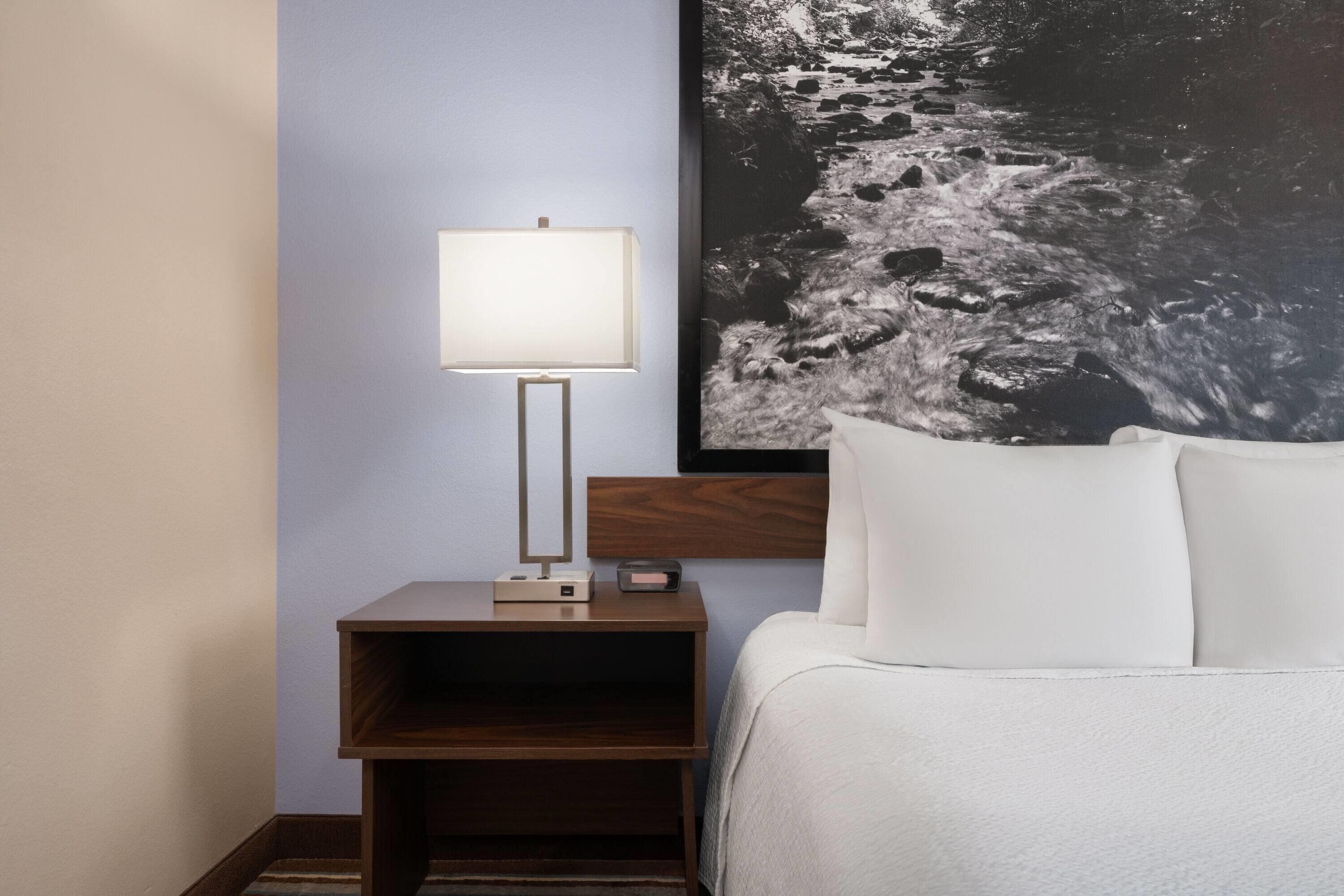 Ein ordentlich bezogenes Bett mit weißen Leinen, ein Nachttisch mit Lampe und ein gerahmtes Bild einer Flusslandschaft an der Wand.