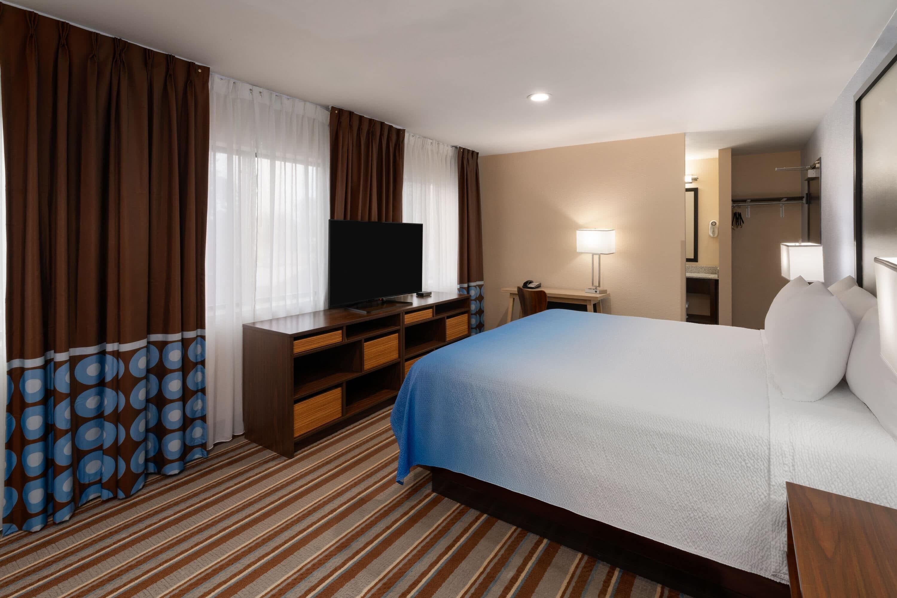 A hotel room with a bed, a TV, a desk, a lamp, and a bathroom visible through an open door.