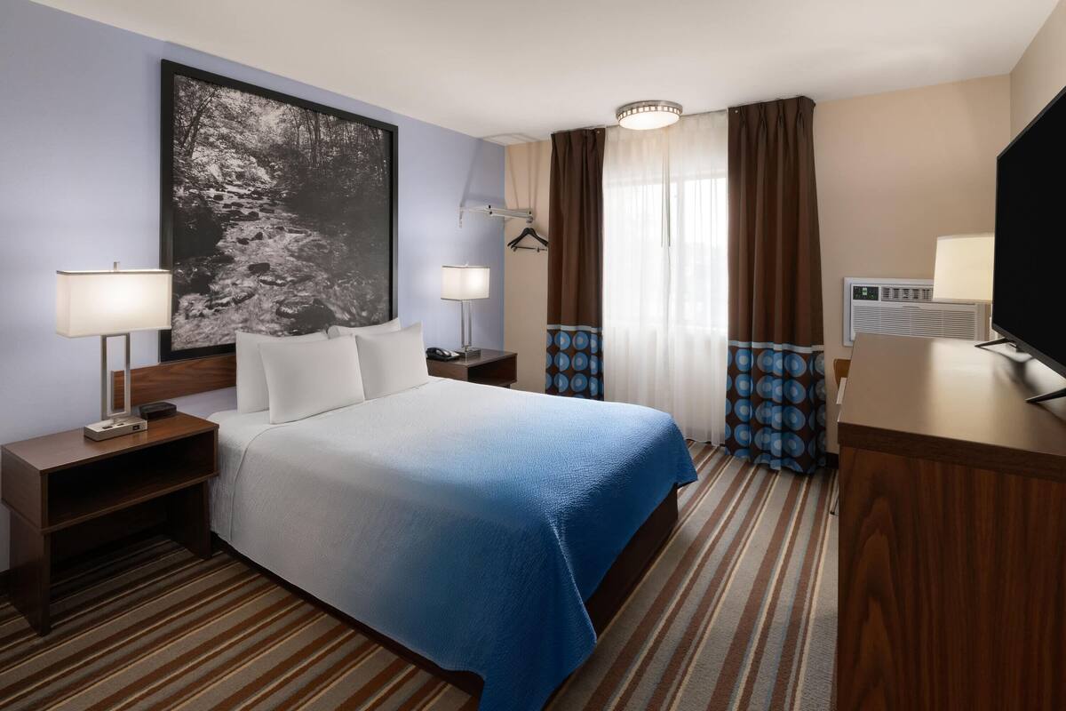 A hotel room with a bed, two bedside tables, a desk, a television, a large painting, and a window with curtains.