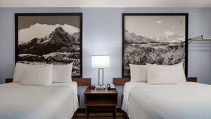 A hotel room with two beds, a nightstand with a lamp, and two large framed black and white mountain landscape pictures on the wall.