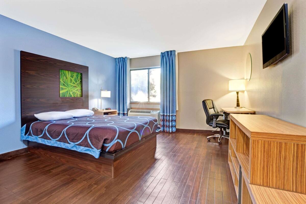 Una camera d'albergo con un letto, una scrivania, una sedia, una TV e una finestra con tende.