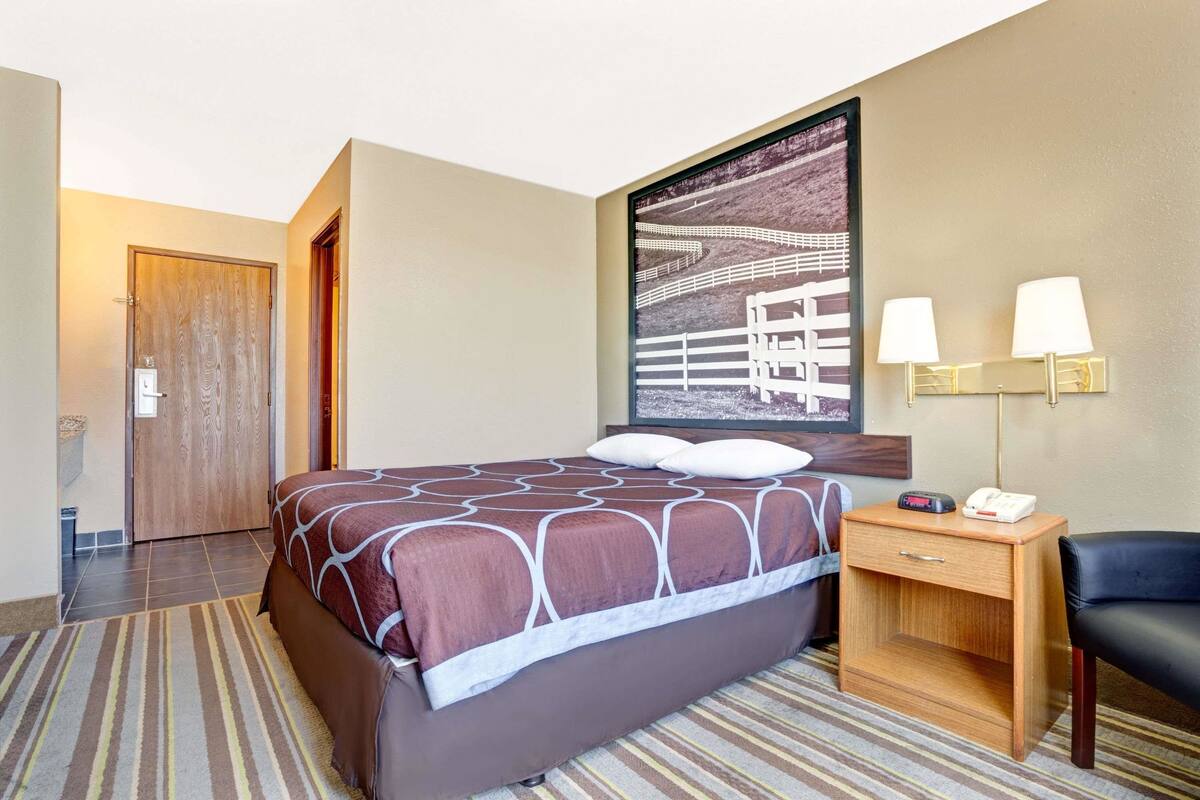 Una camera d'albergo con un letto grande, un comodino con una lampada, una sedia e un quadro appeso al muro.