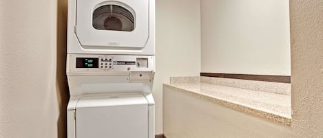 A compact, wall-mounted laundry machine with a digital display and control panel, situated in a corner of a room with a tiled floor and a small built-in bench.