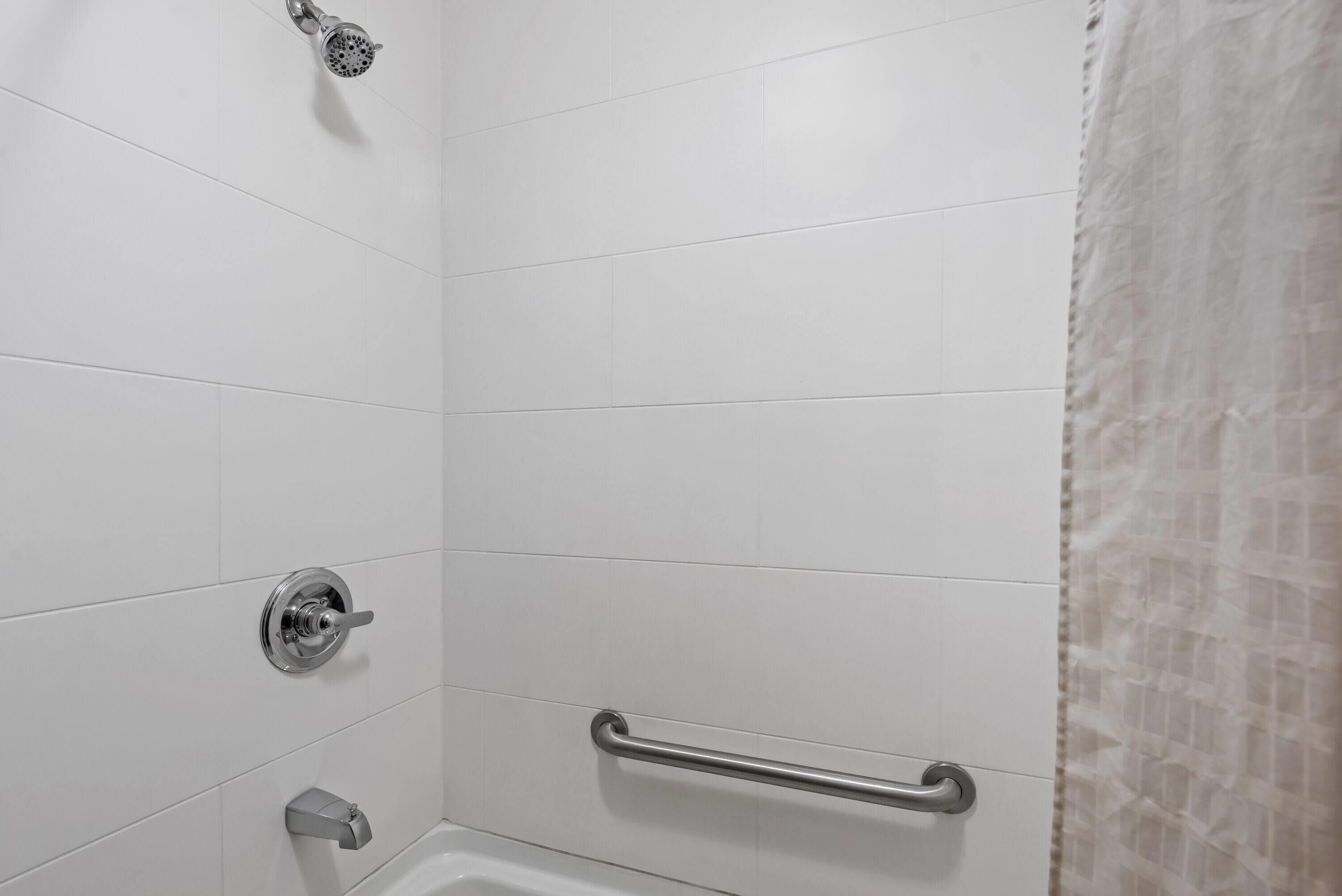 A bathroom with white tiled walls, a showerhead, a grab bar, and a bathtub.