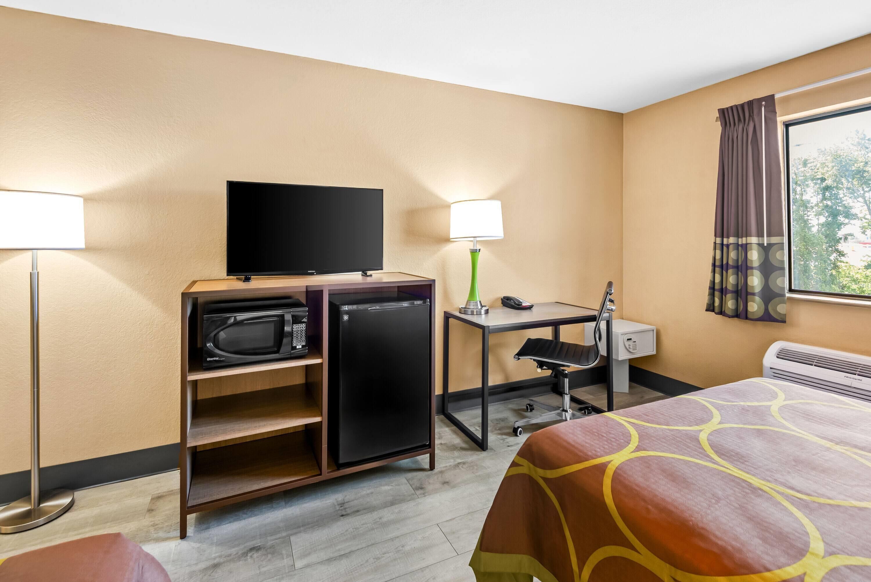 Habitación de hotel con cama, escritorio, computadora, teléfono, televisor, microondas y refrigerador pequeño.