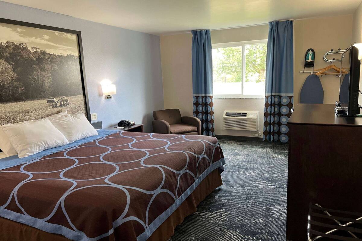 A hotel room with a bed, a chair, a desk, a window with curtains, and a wall-mounted heater.