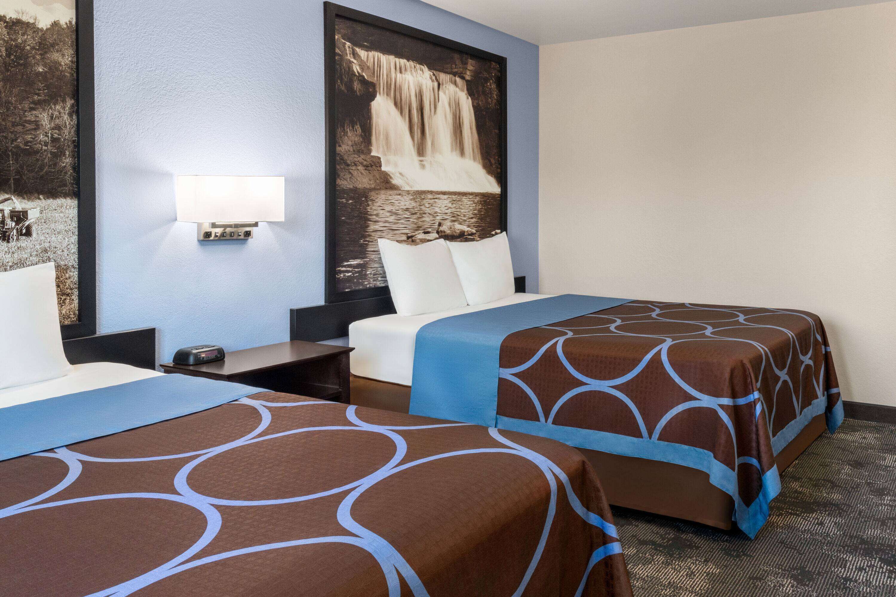 Ein Hotelzimmer mit zwei Betten, einem Nachttisch, einer Wandlampe und einem großen Gemälde eines Wasserfalls.