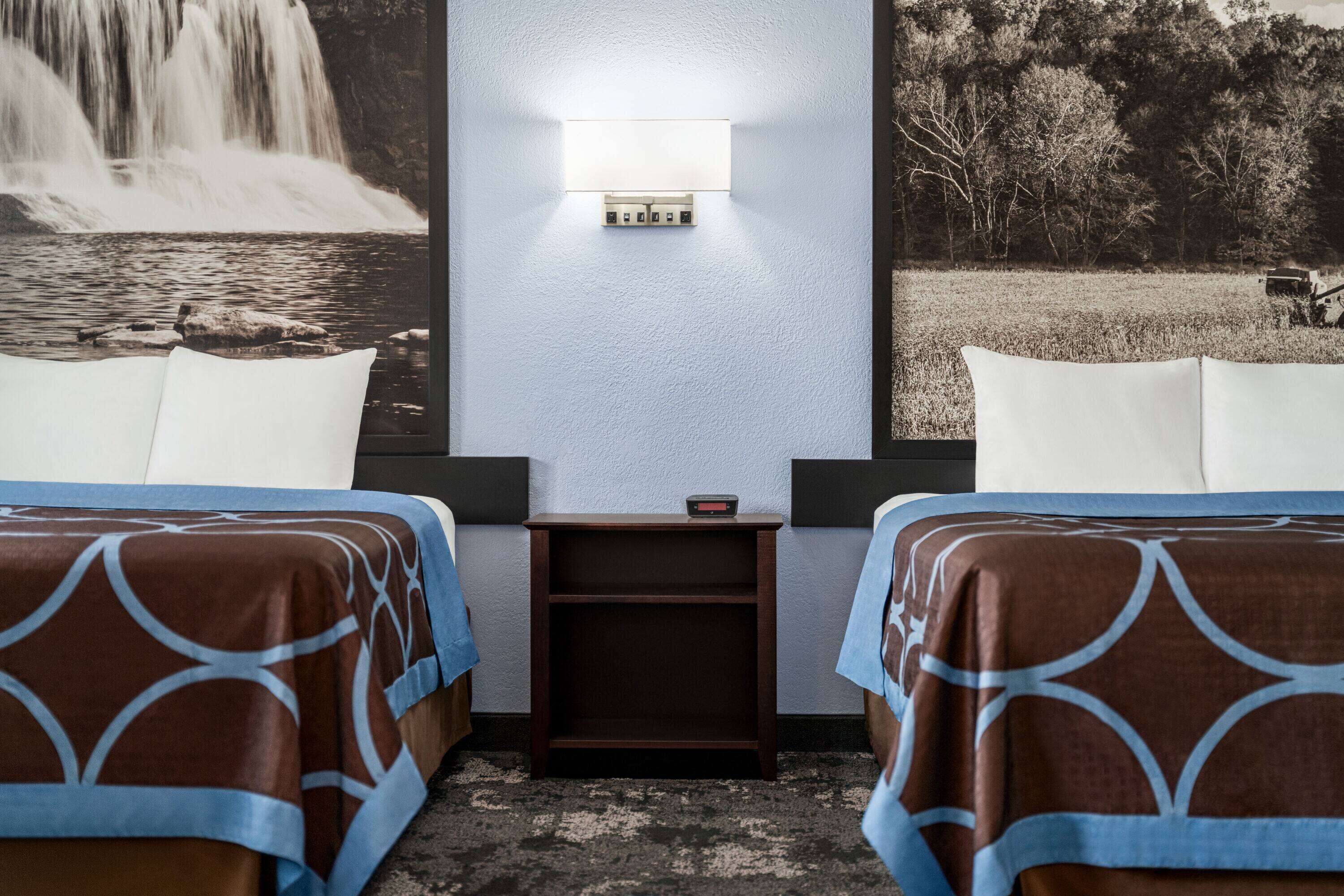 A hotel room with two beds, a nightstand, and a wall-mounted light.