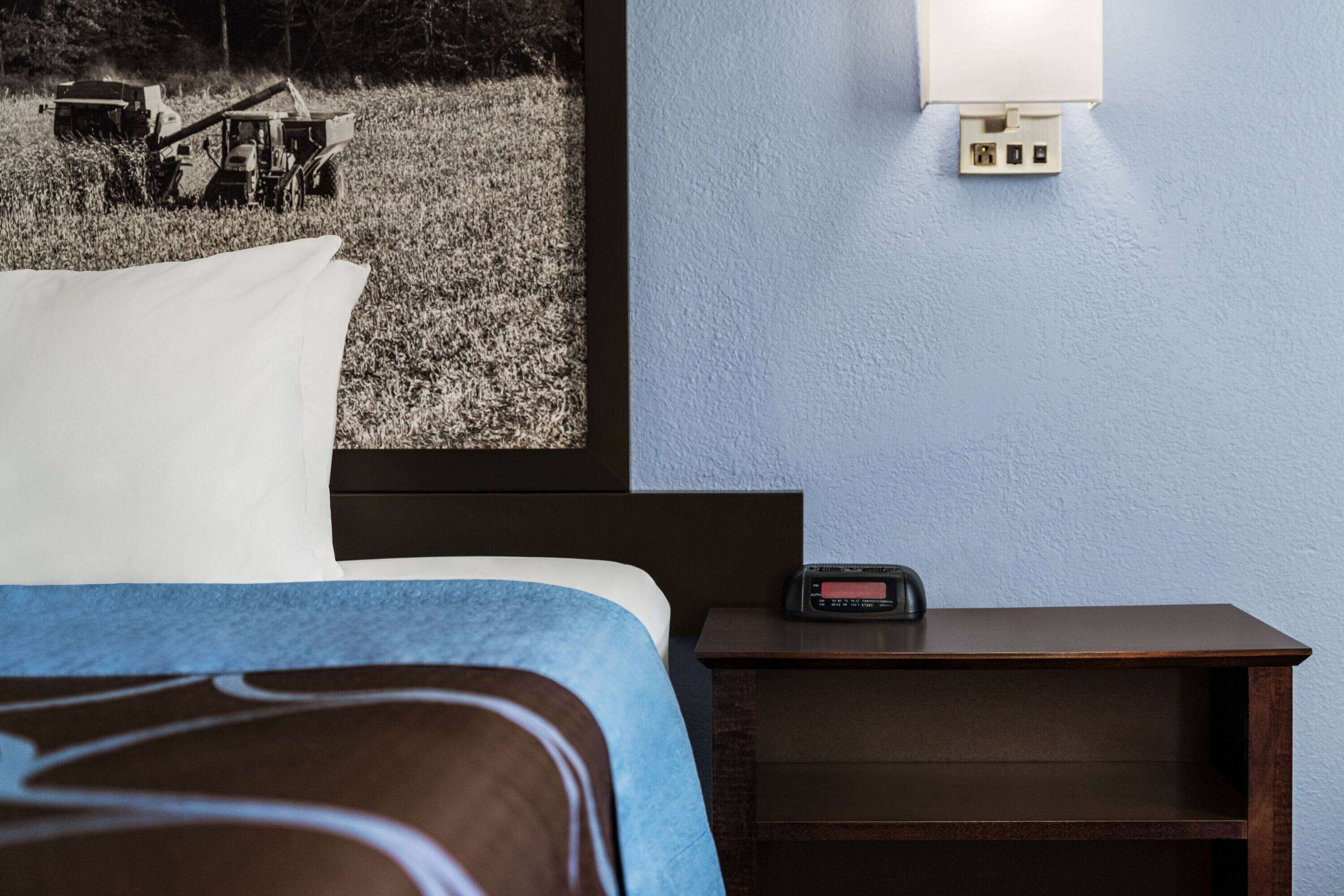 Une chambre d’hôtel avec un lit, une table de chevet dotée d’une horloge numérique, une lampe fixée au mur et une image représentant un tracteur dans un champ.