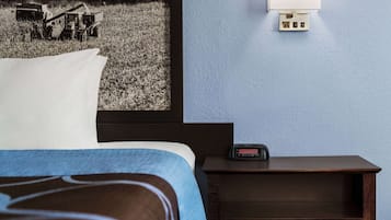 Une chambre d’hôtel avec un lit, une table de chevet dotée d’une horloge numérique, une lampe fixée au mur et une image représentant un tracteur dans un champ.