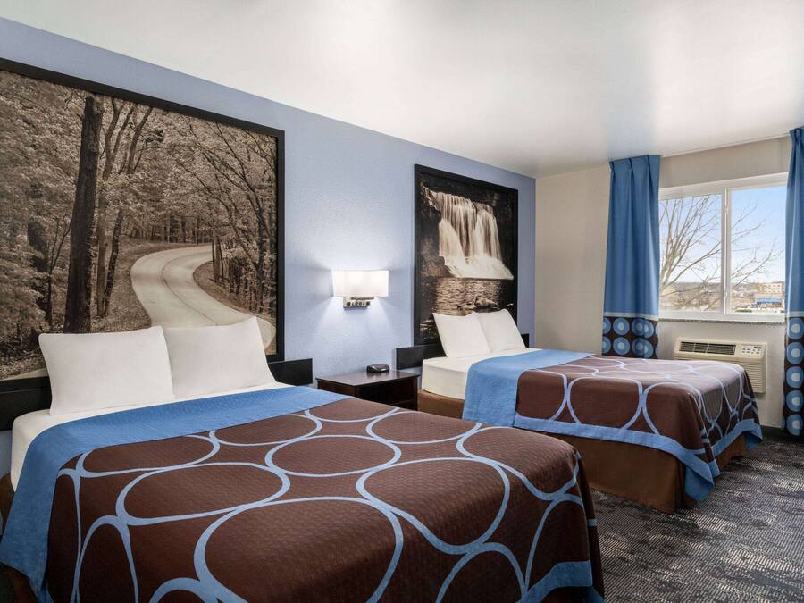 A hotel room with two beds, a large framed picture of a winding road, a nightstand with a lamp, and a window with blue curtains.