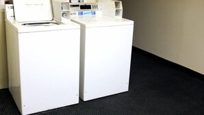 Two white front-loading washing machines in a room with a carpeted floor and a plain wall. - The Lodge at Pensacola (Pensacola)