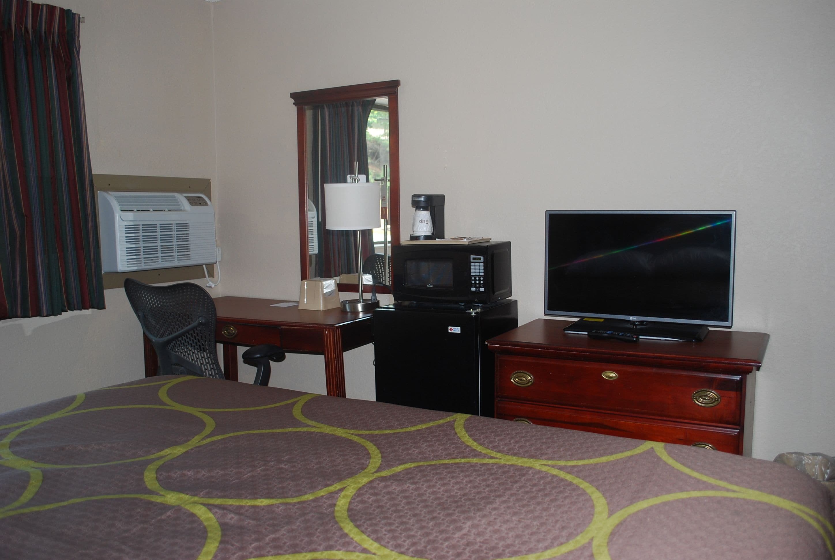 A hotel room with a bed, a desk with a computer, a television, an air conditioner, and a mirror.
