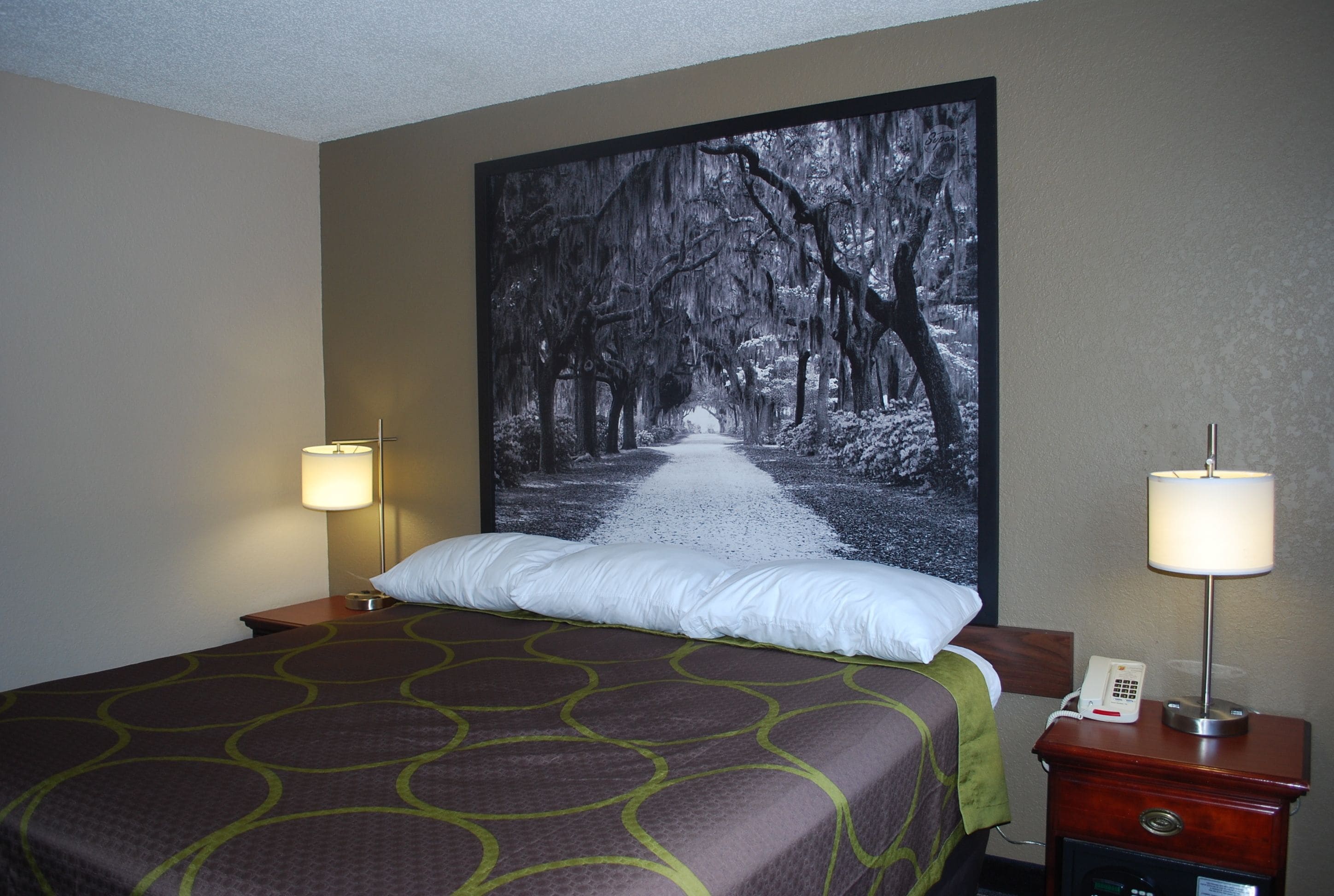 A hotel room with a large bed, two bedside lamps, a nightstand with a phone, and a wall-mounted artwork of a tree-lined path.