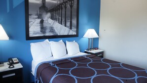 Quarto de hotel com cama, mesa de cabeceira com telefone, luminária e pintura de um cais.