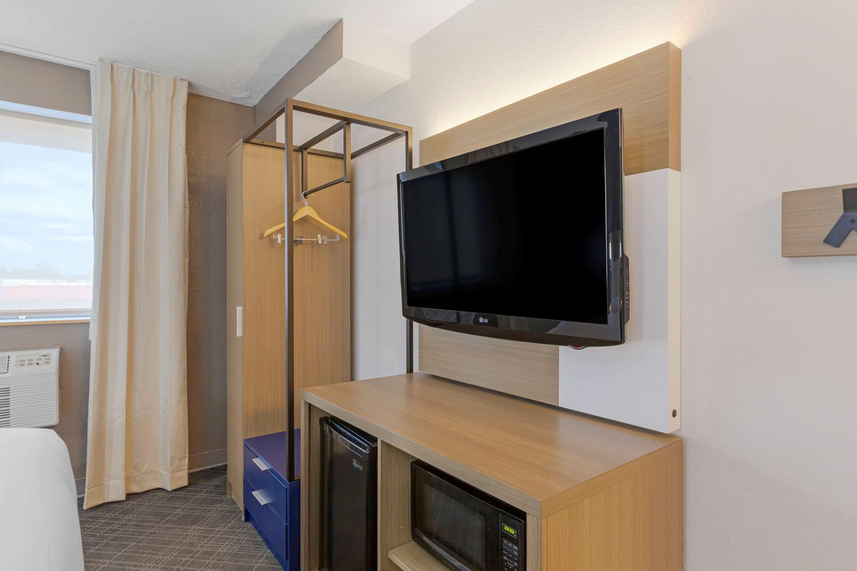 A hotel room with a flat-screen TV mounted on a wooden cabinet, a microwave, and a wardrobe with a hanging rack.