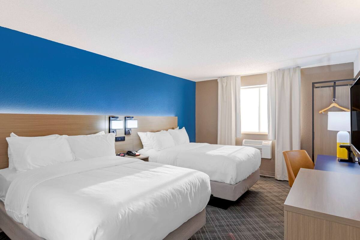 A hotel room with two beds, a desk, a chair, a TV, and a window with curtains.