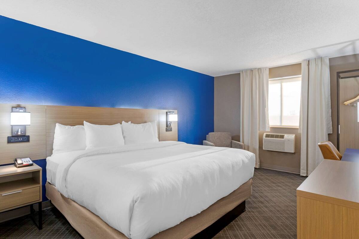 A hotel room with a large bed, bedside tables, a desk, a chair, a window with curtains, and an air conditioning unit.