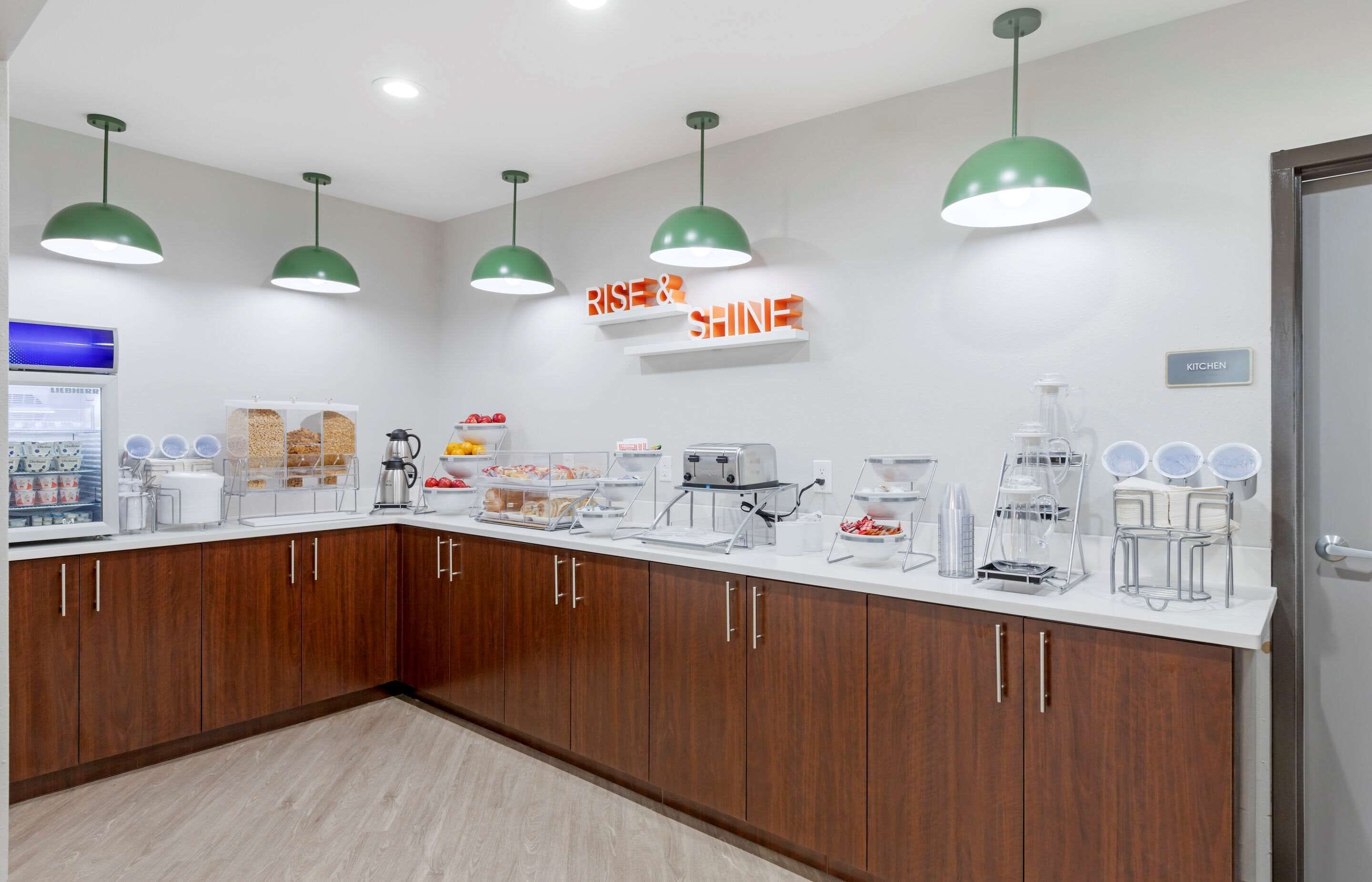 A modern kitchen with wooden cabinets, a white countertop, and green pendant lights. There are various kitchen appliances and a sign that reads "RISE & SHINE".