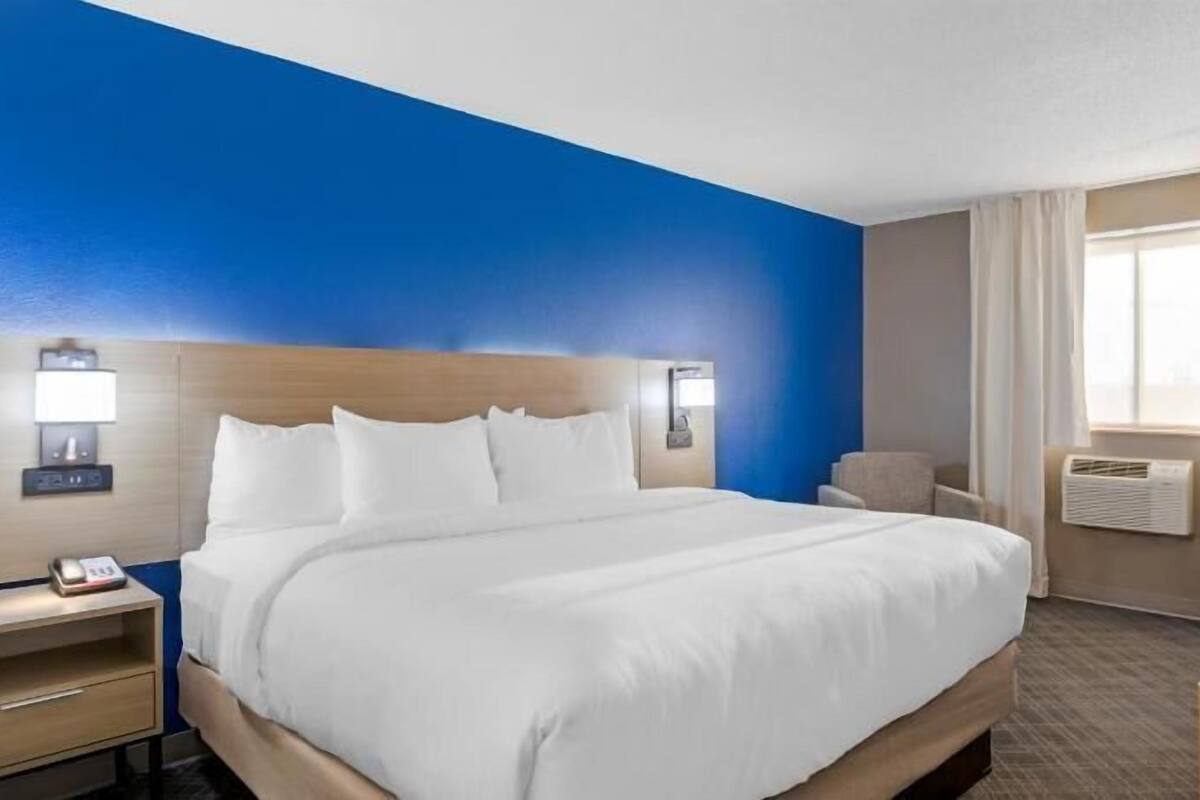 A hotel room with a bed, bedside tables, a desk, an air conditioning unit, and a window with curtains.