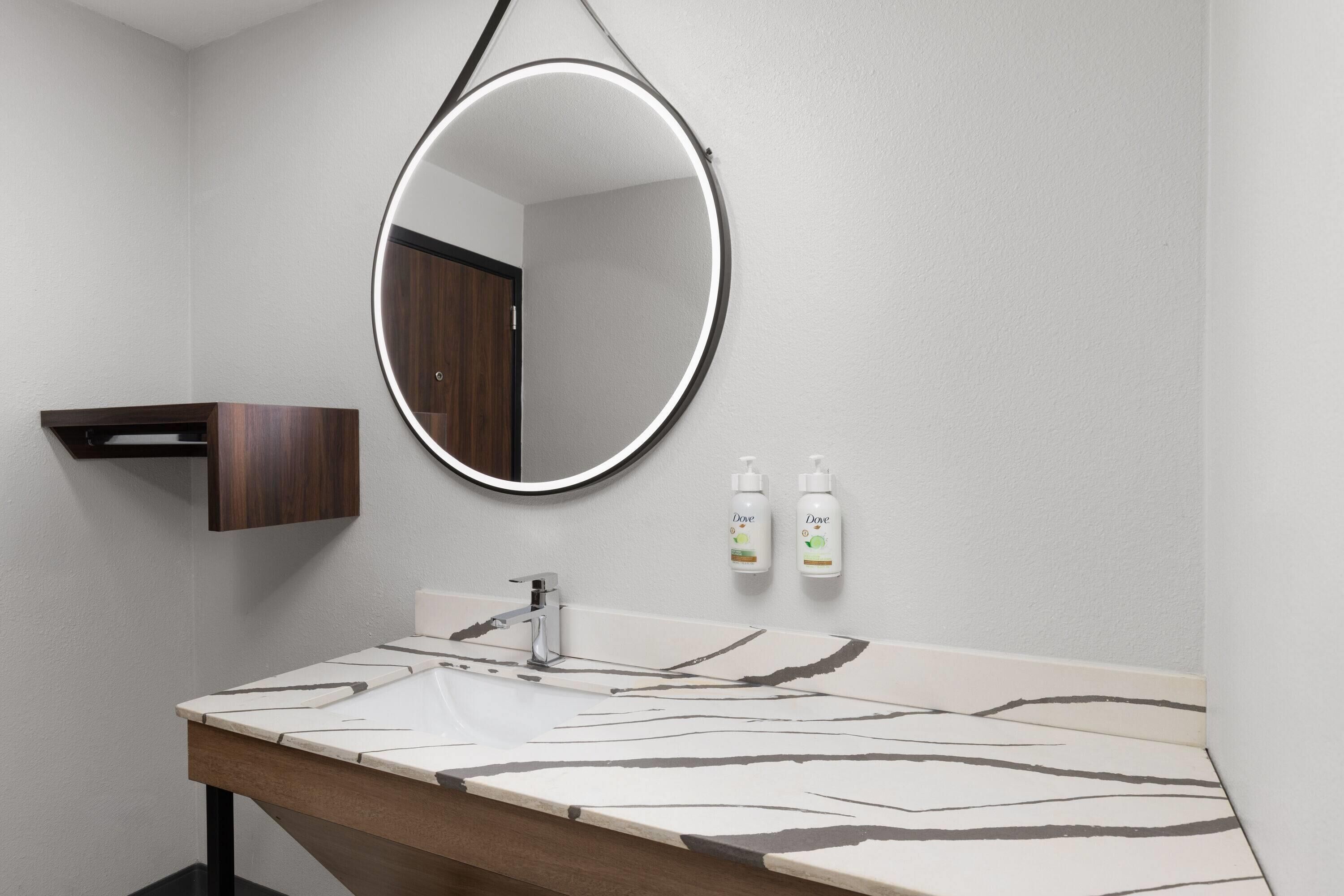 A bathroom with a round mirror, a sink with a marble countertop, and two Dove hand sanitizer bottles.