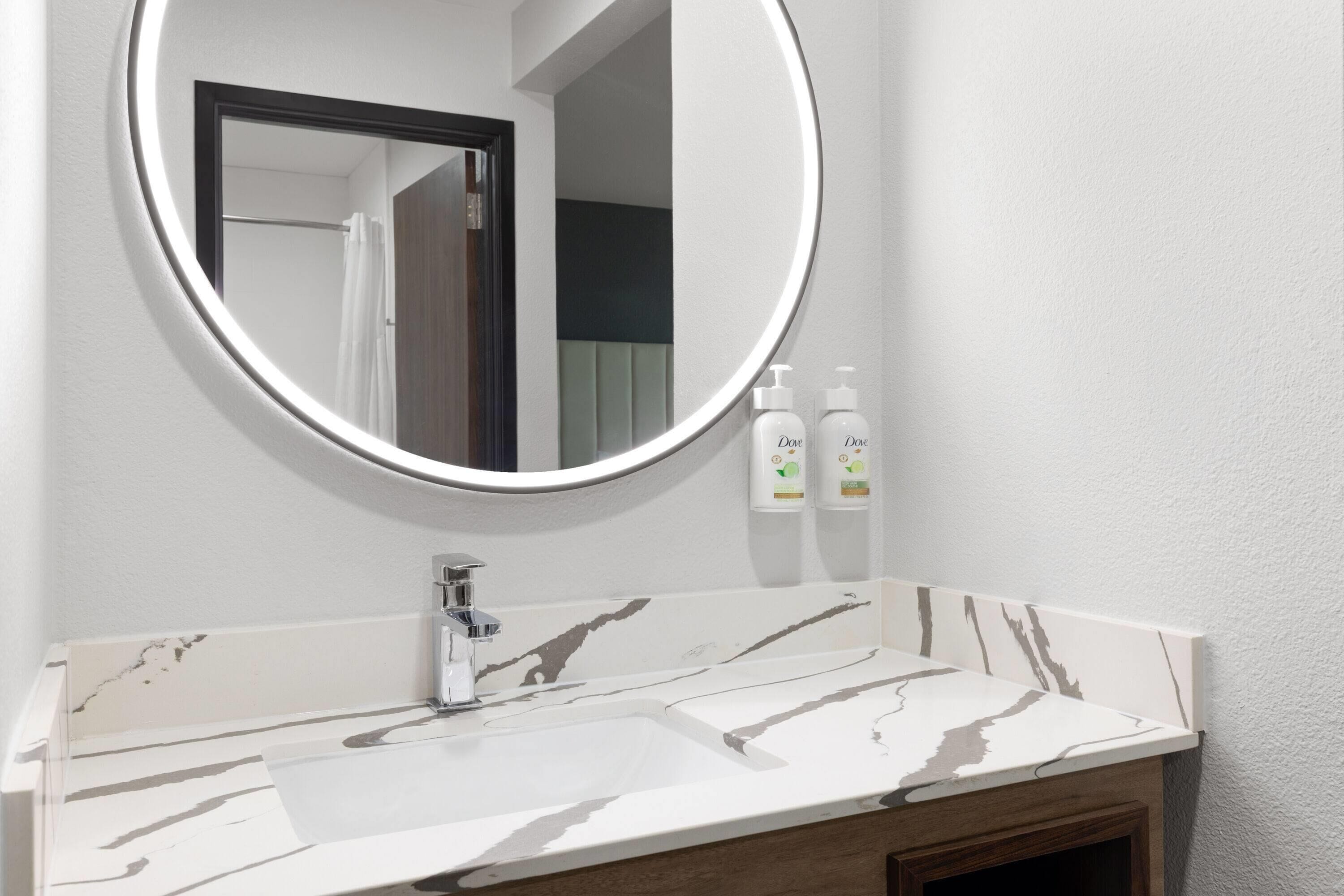 A bathroom with a marble sink, a round mirror, and two Dove hand soap bottles.