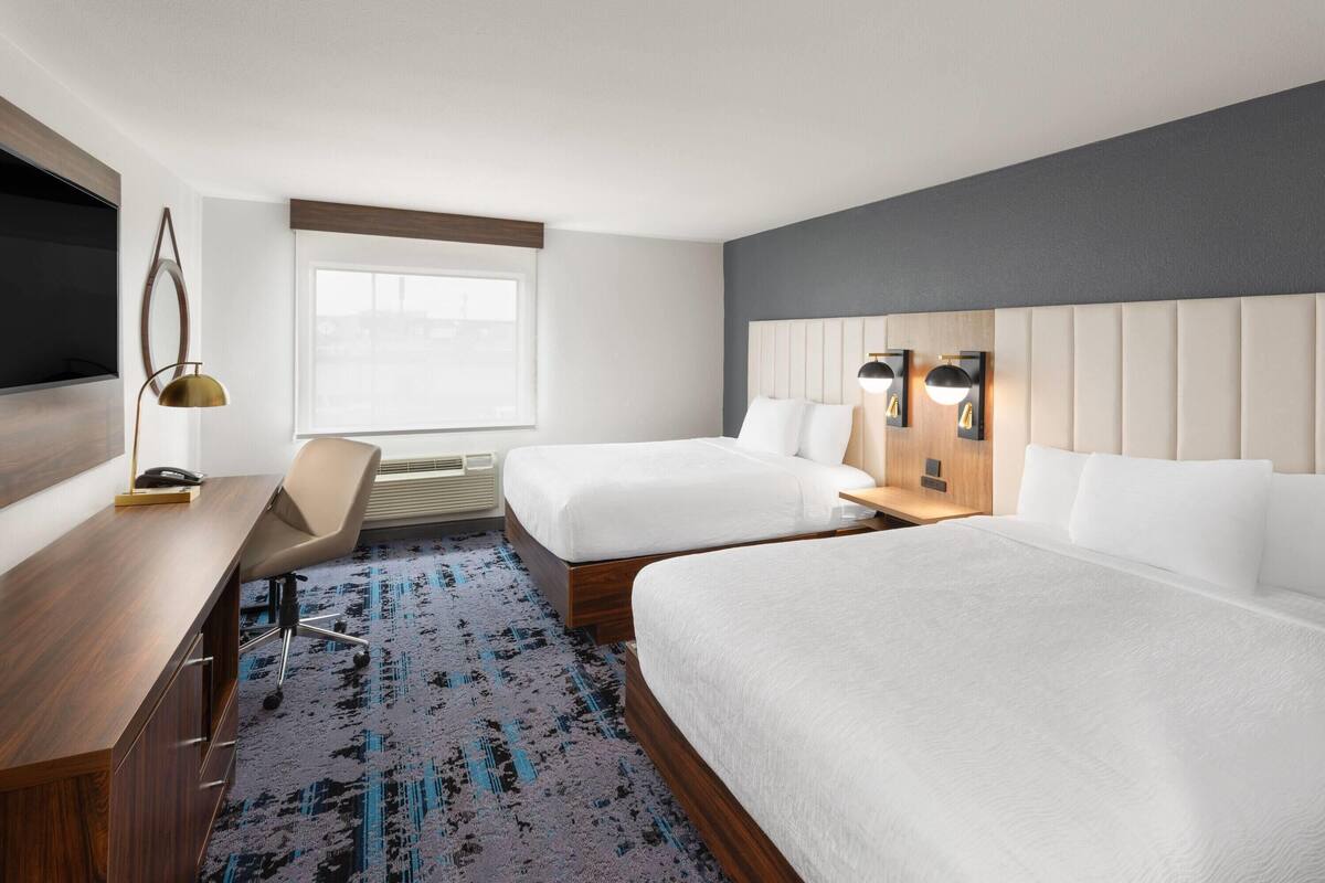 A hotel room with two beds, a desk, a chair, a TV, and a window.