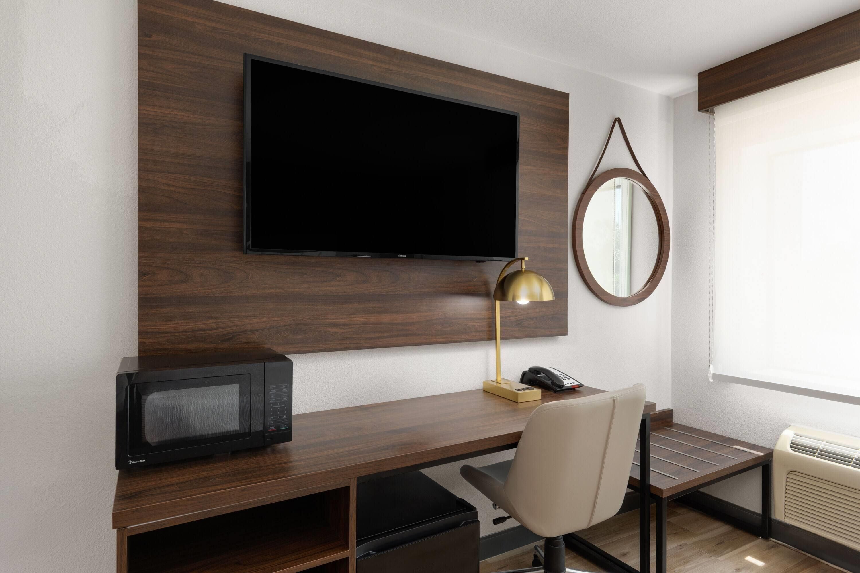 A hotel room with a wooden desk, a flat-screen TV, a desk lamp, a chair, a telephone, and a window with an air conditioning unit.