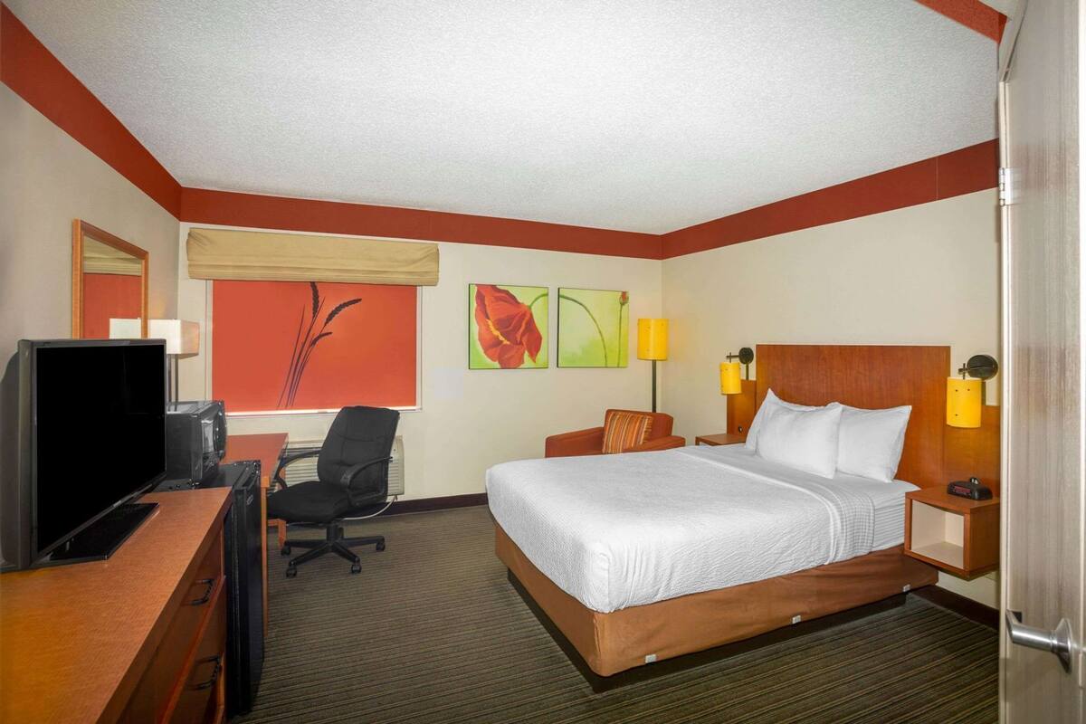A hotel room with a bed, desk, TV, chair, and artwork on the walls.