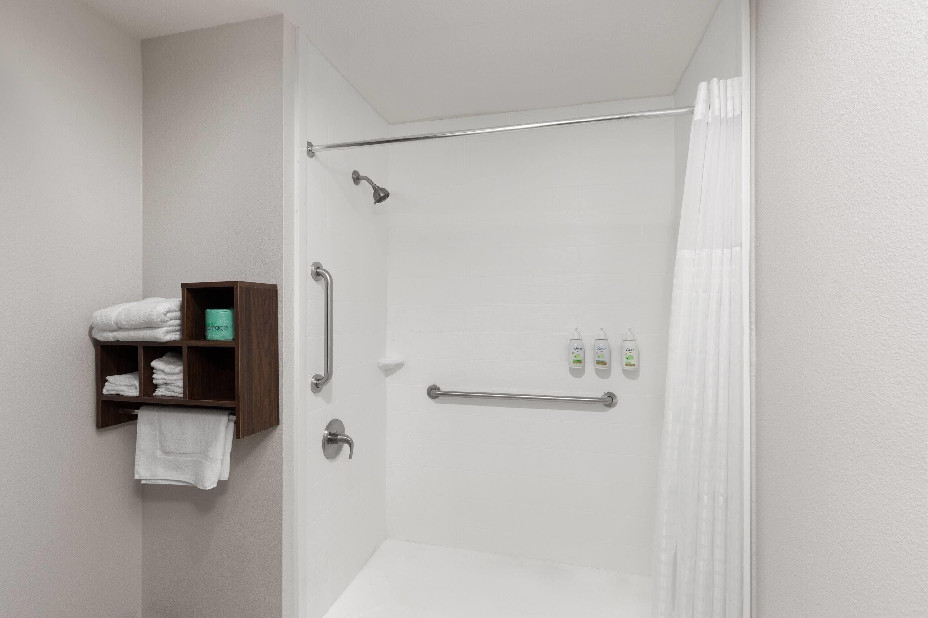 Une douche avec une porte en verre, une étagère en bois avec des serviettes et une barre d’appui.