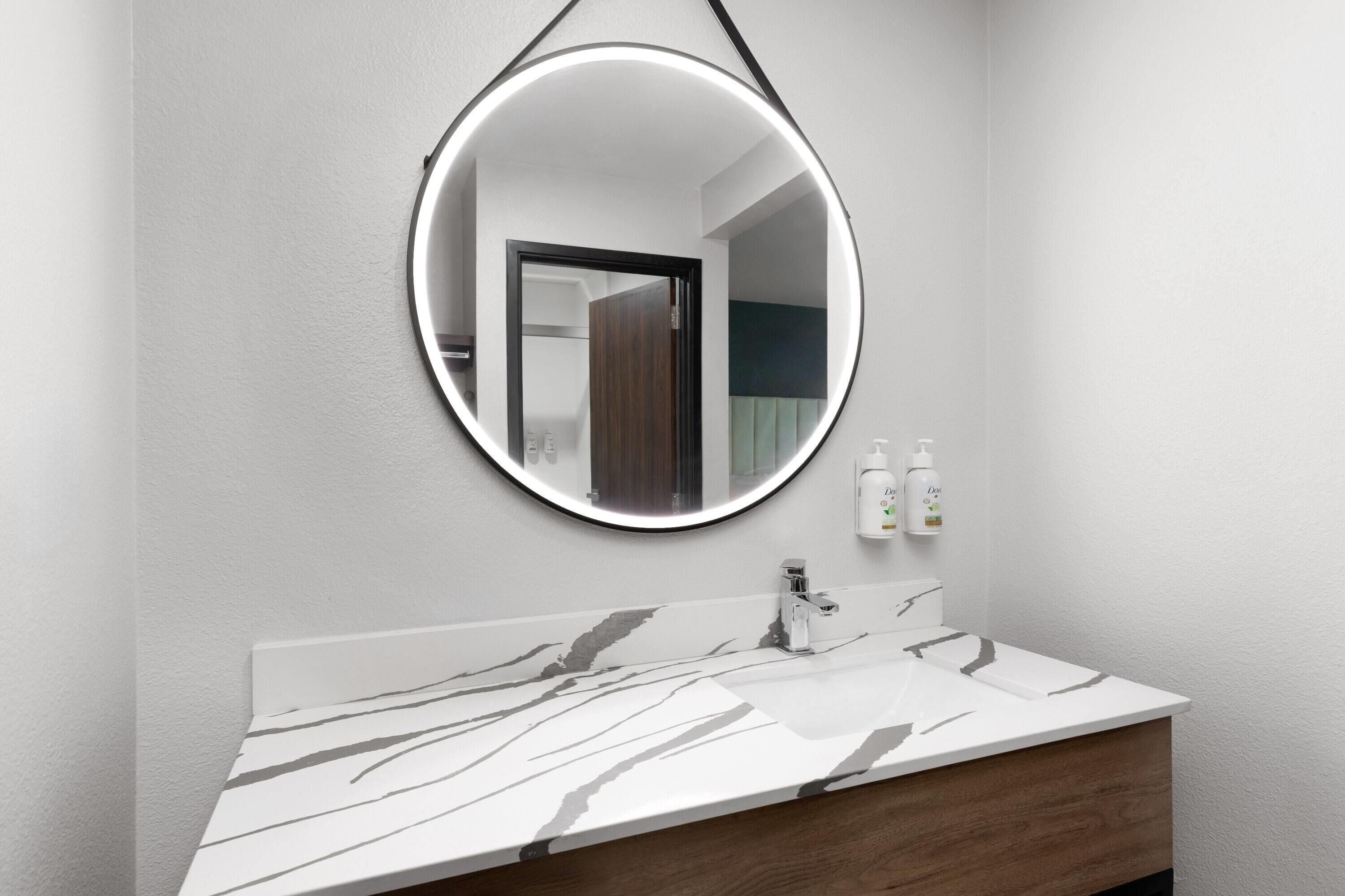 A bathroom with a marble vanity, a round mirror with a light, and two bottles of hand soap on the wall.