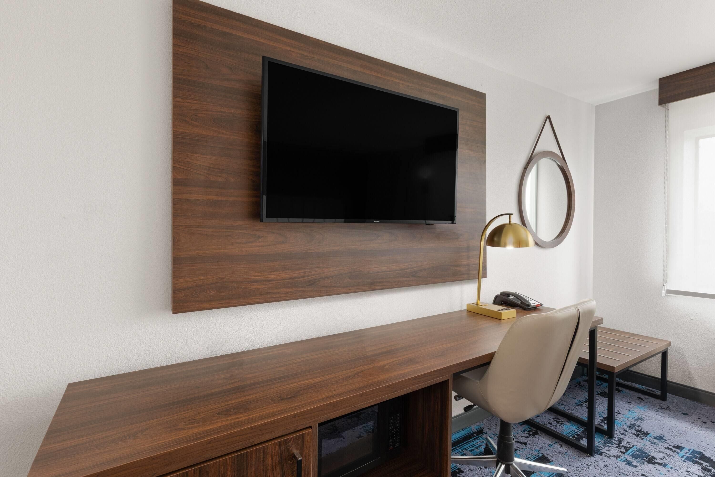 A modern hotel room with a wooden desk, a flat-screen TV mounted on the wall, a desk lamp, and a small mirror.